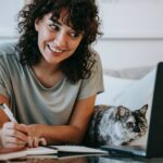 glad woman with cat writing in planner while using laptop