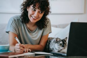 glad woman with cat writing in planner while using laptop